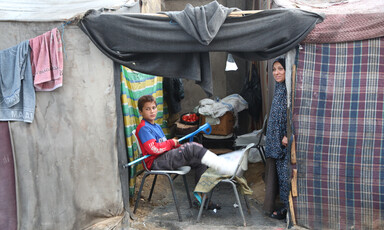 A body with his leg in plastercast sits and a woman stands in a makeshift tent 