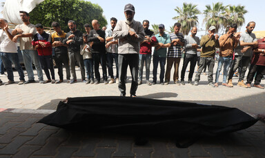 A man stands in front of a dead body in a shroud, with a group of other men behind him