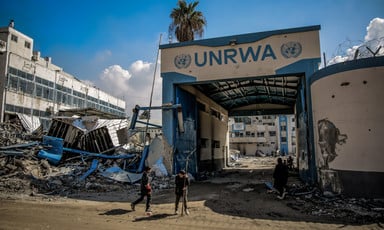 People stand outside gate to UNRWA facility with collapsed structure next to it