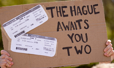 A cardboard protest sign reads "The Hague Awaits You Two" with mock plane tickets for Yoav Gallant and Antony Blinken