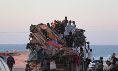 Children sit on top of truck piled high with household objects with sea in background