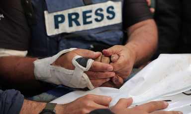 A man wearing a press vest holds the hand of someone wrapped in shroud
