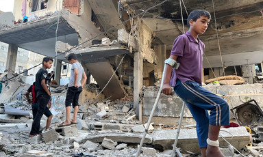 A boy with a bandaged leg uses crutches while walking across rubble in front off bombed-out building
