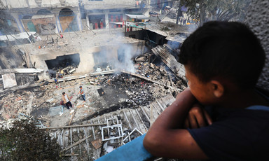 A boy leans over a railing while looking down at smoking debris where a building once stood