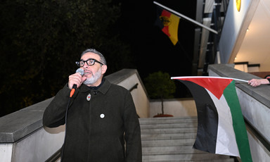 Dr. Abu Sitta speaks in a microphone outside a building with German and Palestinian flags visible behind