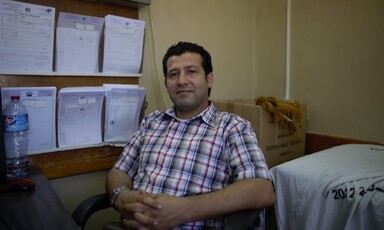 Man gives slight smile to camera while sitting on chair with his hands folding in his lap