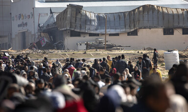 A crowd of people is overlooked by an armored tank in the background