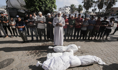 An imam in white leads an outdoor prayer next to two bodies wrapped in white cloth