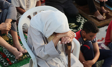 An older person sits, bows their head and leans on a walking stick during prayers in Gaza 