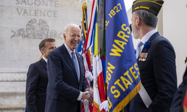 Joe Biden smiles as he shakes hands with a soldier in uniform standing behind a number of flags 