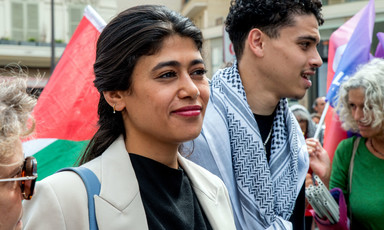 Portrait of the lawmaker Rima Hassan standing in front of a flag and a man wearing a checkered scarf