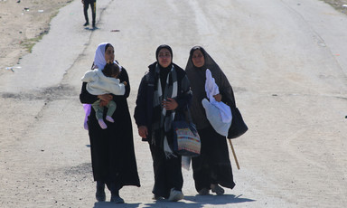 three women walk on a dusty road, one carrying an infant