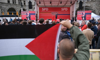 Man holds large Palestinian flag in foreground with man on stage in background