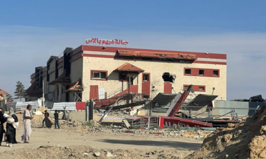 A destroyed wedding hall sits on a dirt road on a bright day with people walking in front