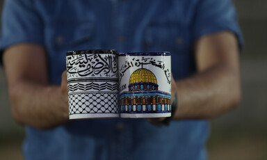 A man holds up two ceramic cups, one with the kefiyeh motif and one depicting al-Aqsa mosque in Jerusalem