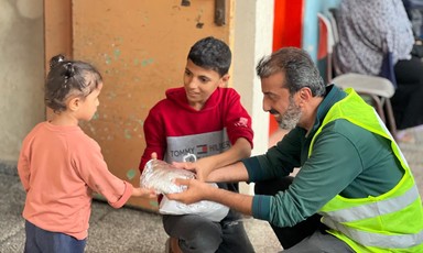 A man wearing a yellow vest hands a wrapped up food parcel to two children in Gaza 