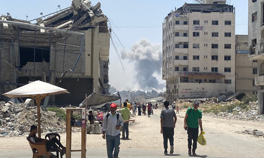 A few people walk beside a badly damaged building with smoke in the background
