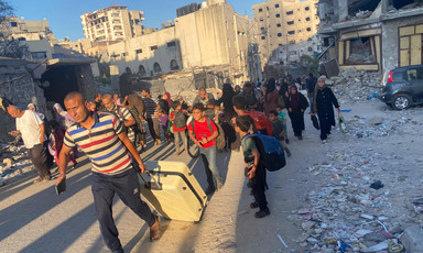 A man in a crowd pulls a suitcase through a neighborhood of destroyed buildings