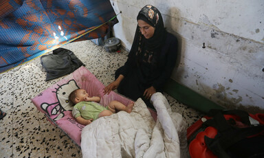 A woman sits against a wall, touching her sleeping infant. 