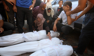A woman sitting on the ground reaches her arm across three shrouded bodies as other mourners stand nearby