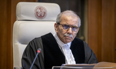 Man wearing judge's robe leans forward while sitting in front of microphone