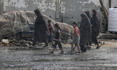 Four women and three children, most wearing sandals, walk along muck-covered road
