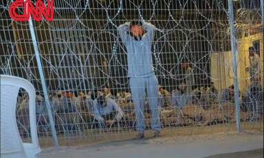 A man wearing a gray tracksuit and with is face mainly covered stands in front of many other men who are seated and behind coils of barbed wire 