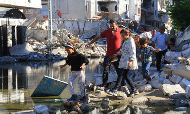 People walk through a pool of sewage water amid the rubble of buildings