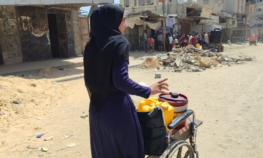 A woman stands behind a wheelchair containing water containers 