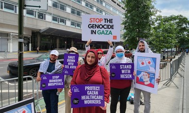 Woman holds sign reading "Protect Gazan healthcare workers" while standing infant of five others holding signs and wearing keffiyehs