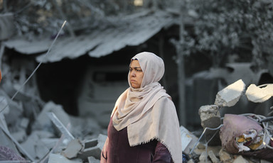 A woman stands in front of the rubble of a house