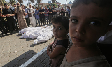 Two young children look at camera while standing next to four shrouded bodies during funeral