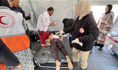 A woman wearing a hoodie with a Red Crescent logo stands over a boy lying face down on a stretcher as other medics work around them