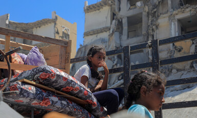 Two girls sit on top of mattresses with destroyed buildings in background