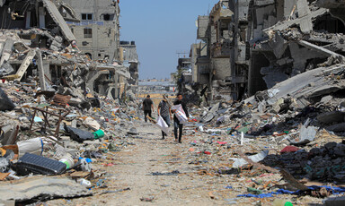 Three men walk in the middle of the road surrounded on either side by destroyed buildings spilling out their household contents