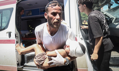 A man carries an injured child out of an ambulance with an older child and woman in the background