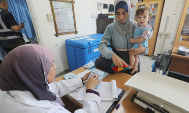 A woman holds a baby who looks towards the camera while standing in front of health clinic examination table