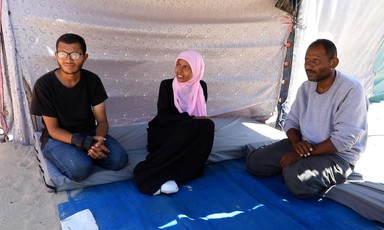 Sitting on a blue cloth under a shade are a young man, a young woman and an older man