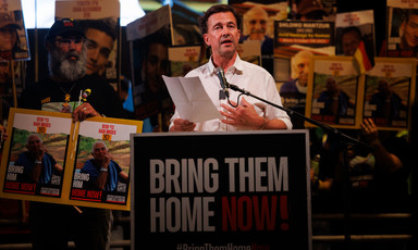 Steffen Seibert stands at a lectern surrounded by people carrying signs supporting Israelis held in Gaza