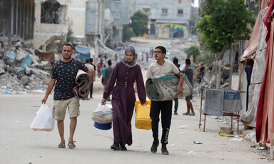 Three people walk with bags and blankets on a backdrop of ruined buildings