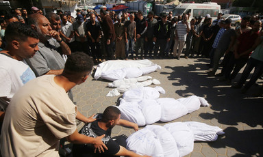 A group of mourners surround six corpses of various sizes wrapped in white shrouds