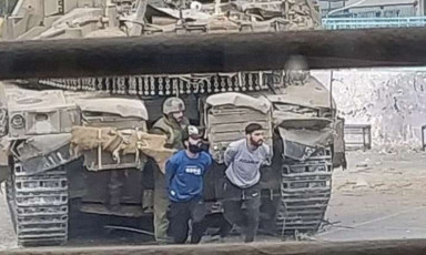 Two men with arms behind their back walk in front of an Israeli tank