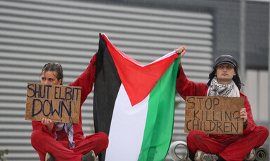 Protesters wearing read hold a Palestinian flag and cardboard signs with slogans opposing the activities of the Israeli weapons maker Elbit Systems 