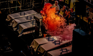 A person holds a flare emitting fiery-looking orange smoke while standing next to coffins draped in Israeli flags