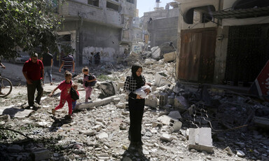 Palestinian children and adults, including a woman holding an infant, step across a rubble-strewn street