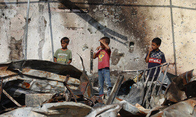 Three children stand on the charred and twisted ruins of shelters hit by an Israeli airstrike.