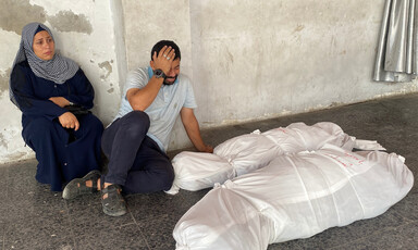 A man and a woman mourn next to two bodies wrapped in white shrouds