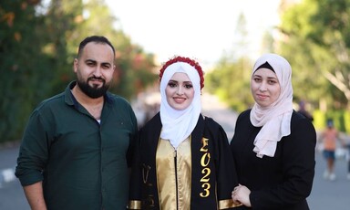 In the daytime, three people smile for the camera, one in graduation gown