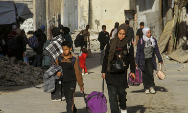 People, including children, carrying their belongings walk down a street strewn with rubble