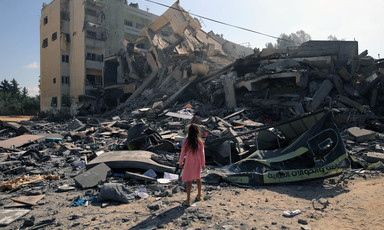 A girl in a pink dress is seen from the back while standing in front of a destroyed building